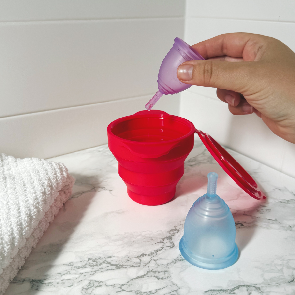 Person placing a purple Ruby Cup menstrual cup into a red steriliser on a marble surface.