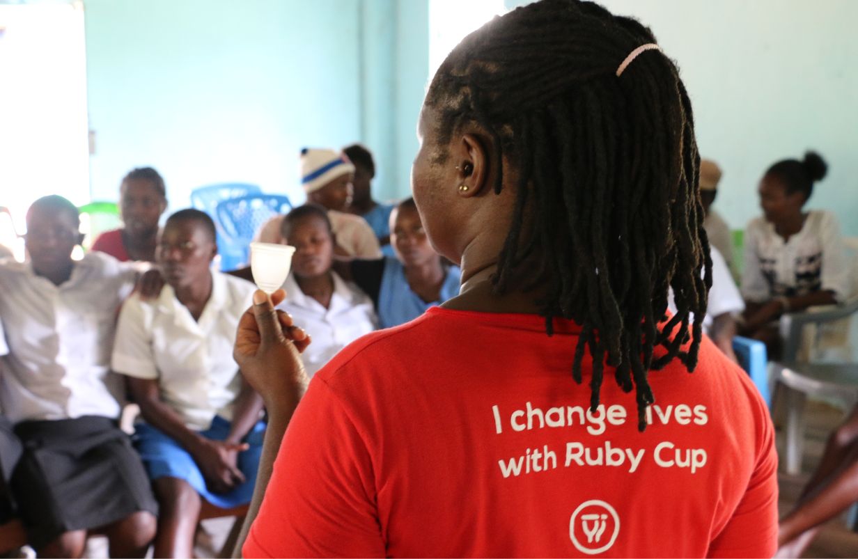 Person in a red shirt with 'I change lives with Ruby Cup' text, facing a group of people and holding a Ruby Cup menstrual cup.