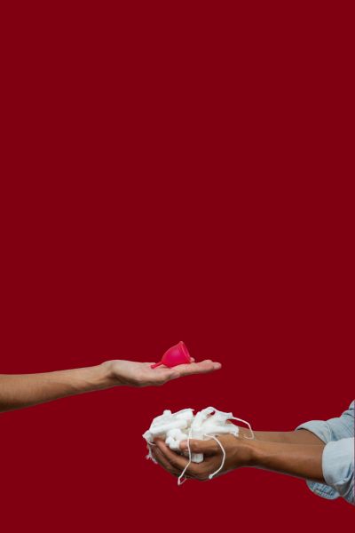 Two hands: one holding a red Ruby Cup menstrual cup, the other holding disposable tampons.